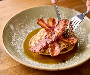 A knife and fork cutting into a stack on pancakes topped with streaky bacon and maple syrup
