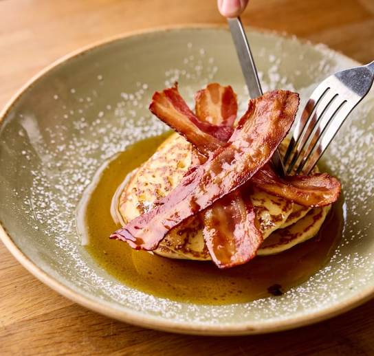 A knife and fork cutting into a stack on pancakes topped with streaky bacon and maple syrup