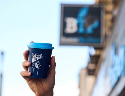 A hand holding a blue reusable coffee cup with the words 'Be an everyday hero' in white written across it