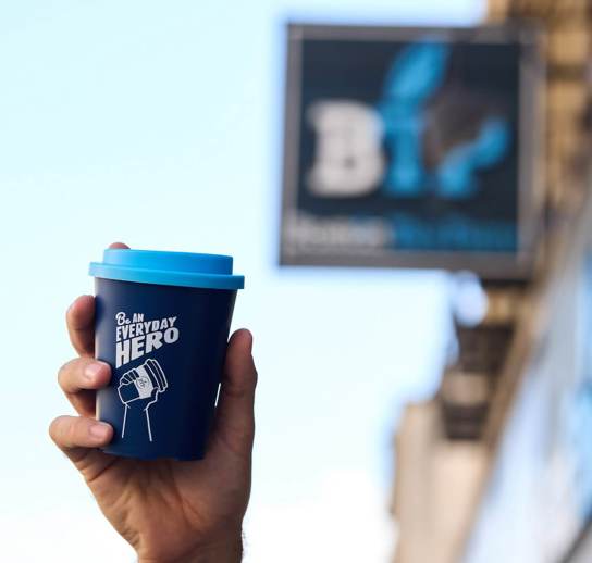 A hand holding a blue reusable coffee cup with the words 'Be an everyday hero' in white written across it